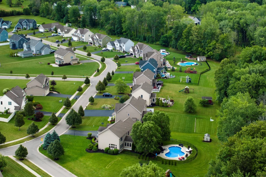 Aerial view of a suburban residential neighborhood with neatly arranged houses, green lawns, tree-lined streets, and several backyard pools.