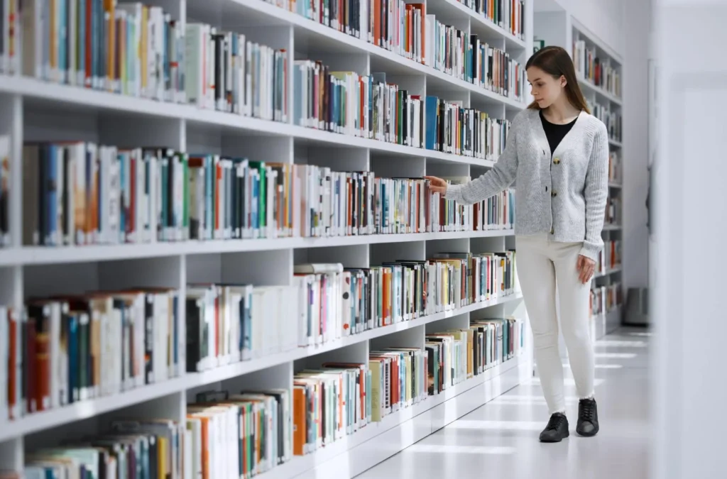 Woman browsing books along a bright white library shelf, reaching for a title in a clean, well-organized space that reflects safe, well-managed library security and access