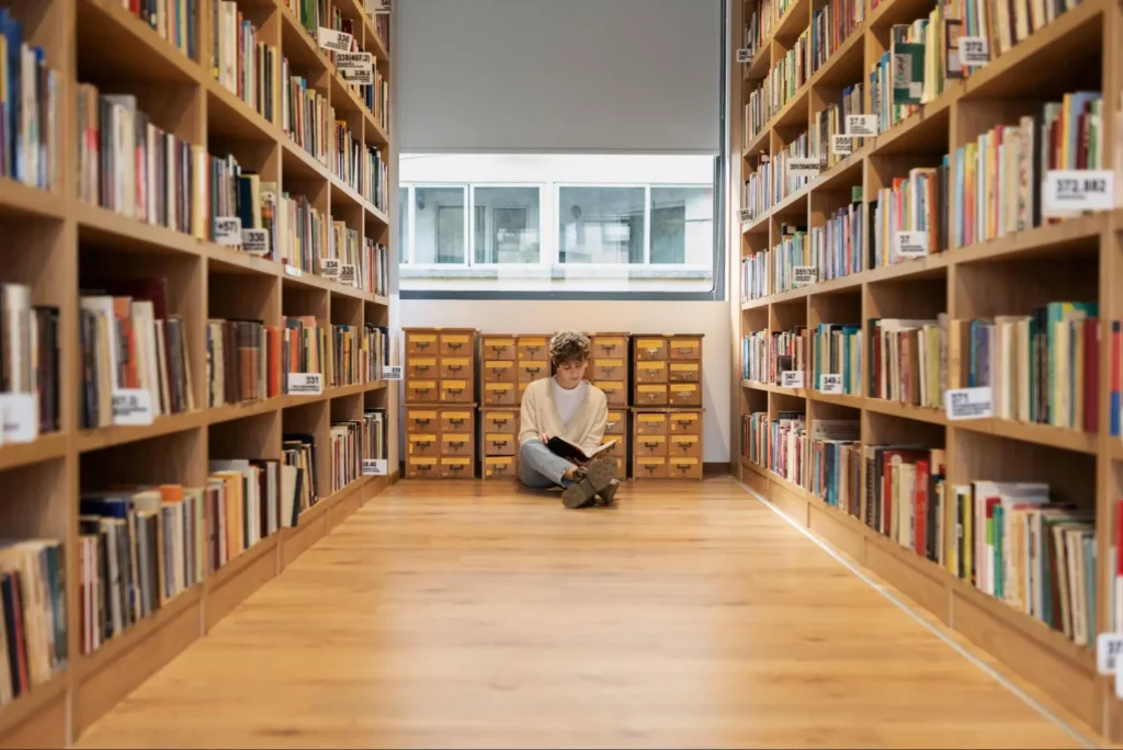 Young student sitting cross-legged on the floor of a library aisle reading a book, surrounded by tall wooden bookshelves and a vintage card catalog under natural window light